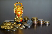 Figure Of The Golden Buddha On The Table. Next To It Is A Stack Of Coins. Symbol Of Wealth And Prosperity.