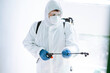 © maxbelchenko - Man in Hazmat suit, protective gloves and goggles use sprayer equipment disinfect the entrance of a residential building. Covid-2019.