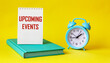 Conceptual message UPCOMING EVENTS in notepad and on yellow background, next to alarm clock and green diary. The best view of the modern workplace.