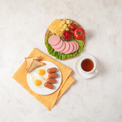 Delicious breakfast - two eggs with fried sausages and toast bread, cap of tea, wooden cutting board with salad, tomatoes, sausage and cheese on white textured background. Copy space.