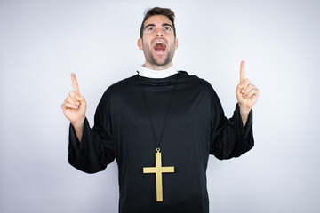 Wall Mural - Young hispanic man wearing priest uniform standing over white background amazed and surprised looking at the camera and pointing up with fingers and raised arms