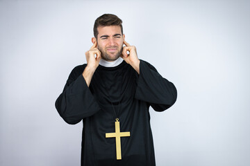 Wall Mural - Young hispanic man wearing priest uniform standing over white background covering ears with fingers with annoyed expression for the noise of loud music. Deaf concept.