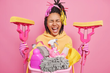 Wall Mural - Home cleaning and housekeeping concept. Dark skinned housewife exclaims loudly holds two mops dressed in yellow sweatshirt poses near basket of dirty laundry isolated over pink studio background.