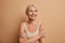 Attractive Positive Wrinkled Fifty Years Old Woman Looks Gladfully Above Keeps Arms Foded Has Well Cared Complexion Healthy Skin White Teeth Isolated Over Brown Background. Beauty And Age Concept