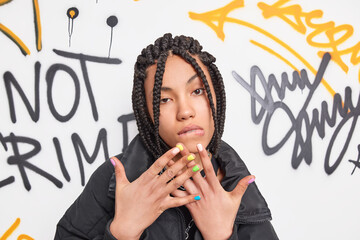 Poster - Self confindent cool teenager looks seriously at camera raises hands bites lips has dreadlocks hairstyle wears fashionable clothes poses against graffiti wall. Youth fashion teenage lifestyle concept