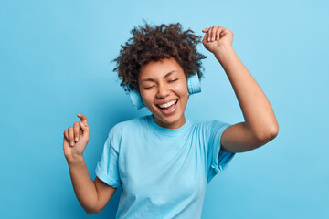 Wall Mural - Joyful relaxed African American girl enjoys favorite playlist listens music via wreless headphones raises arms dressed casually isolated over blue background. People hobby and lifestyle concept