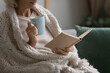 © fizkes - Old retired 70s lady reading book at home, drinking hot tea, resting on sofa in cozy living room. Mature reader wrapped in knitted blanket relaxing with novel over cup of coffee or chocolate. Close up