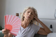© fizkes - Mature senior woman exhausted with heat, swelter, summer weather, waving hand fan at home, cooling in too hot air. Tired unhappy elderly 60s lady suffering from hormone imbalance menopause symptoms