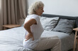 © fizkes - Tired upset elderly woman feeling strong lower backpain after sleeping in incorrect posture on uncomfortable mattress. Senior 60s lady with closed eyes sitting on bed, holding painful lumber muscles