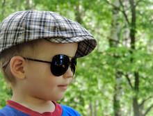 A Four Year Old Boy In A Checkered Cap And Sunglasses Looks Into The Distance Against The Background Of A Blurry Bokeh Forest.