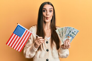 Wall Mural - Beautiful brunette young woman holding united states flag and dollars making fish face with mouth and squinting eyes, crazy and comical.