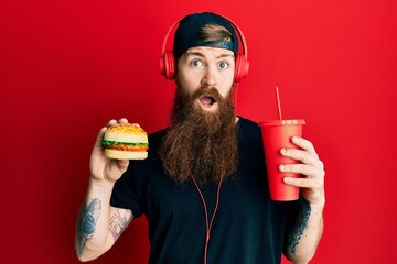 Wall Mural - Redhead man with long beard eating a tasty classic burger and drinking soda clueless and confused expression. doubt concept.