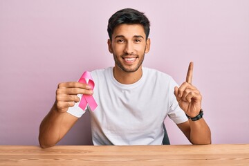 Young handsome man holding pink cancer ribbon surprised with an idea or question pointing finger with happy face, number one