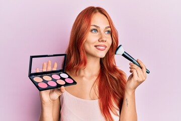 Poster - Young redhead woman holding makeup and brush smiling looking to the side and staring away thinking.