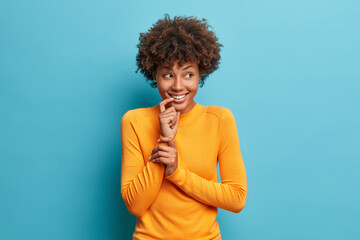 Good looking cheerful Afro American woman smiles gently wears casual long sleeved jumper looks joyful aside poses against blue background. Satisfied ethnic young female model stands thoughtful