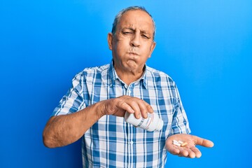 Poster - Senior hispanic man holding pills puffing cheeks with funny face. mouth inflated with air, catching air.