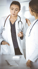 Wall Mural - Two female physicians are discussing medical therapy, while standing at the table in a sunny clinic office. Doctors at work. Teamwork in medicine