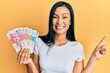 © Krakenimages.com - Beautiful hispanic woman holding hong kong dollars banknotes smiling happy pointing with hand and finger to the side