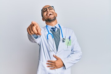 Wall Mural - Hispanic adult man wearing doctor uniform and stethoscope laughing at you, pointing finger to the camera with hand over body, shame expression