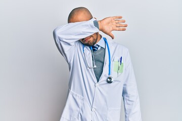 Sticker - Hispanic adult man wearing doctor uniform and stethoscope covering eyes with arm, looking serious and sad. sightless, hiding and rejection concept