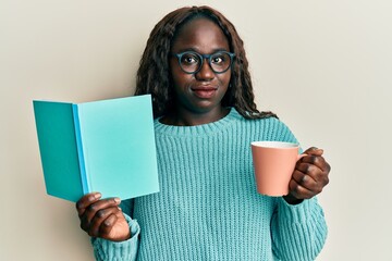 Poster - African young woman reading a book and drinking a cup of coffee relaxed with serious expression on face. simple and natural looking at the camera.