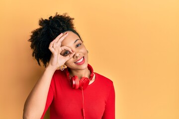 Wall Mural - Young african american girl listening to music using headphones smiling happy doing ok sign with hand on eye looking through fingers