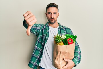 Wall Mural - Handsome man with beard holding paper bag with groceries looking unhappy and angry showing rejection and negative with thumbs down gesture. bad expression.