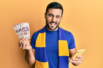 Poster - Young hispanic man football supporter using smartphone holding mexican pesos banknotes winking looking at the camera with sexy expression, cheerful and happy face.