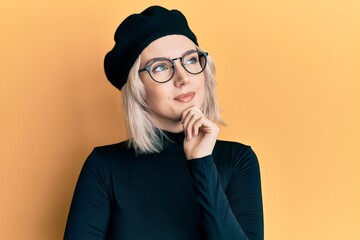 Poster - Young blonde girl wearing french look with beret looking confident at the camera with smile with crossed arms and hand raised on chin. thinking positive.