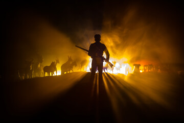 silhouette of a man (hunter) with rifle standing against group of animals in colorful dark backlight