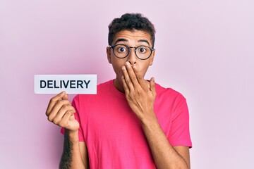 Poster - Young handsome african american man holding delivery word covering mouth with hand, shocked and afraid for mistake. surprised expression