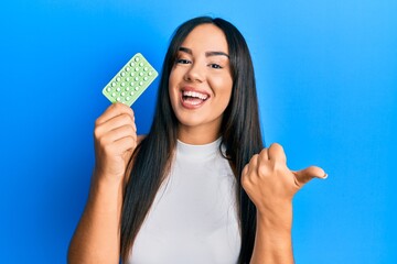 Young beautiful hispanic girl holding birth control pills pointing thumb up to the side smiling happy with open mouth