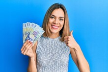 Young Caucasian Blonde Woman Holding 100 Romanian Leu Banknotes Smiling Happy And Positive, Thumb Up Doing Excellent And Approval Sign
