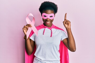 Wall Mural - Young african american girl wearing superhero costume holding pink cancer ribbon surprised with an idea or question pointing finger with happy face, number one