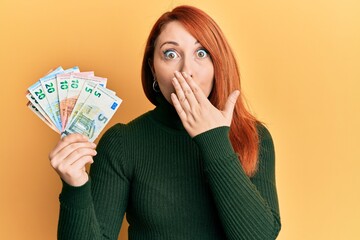 Wall Mural - Beautiful redhead woman holding euro banknotes covering mouth with hand, shocked and afraid for mistake. surprised expression