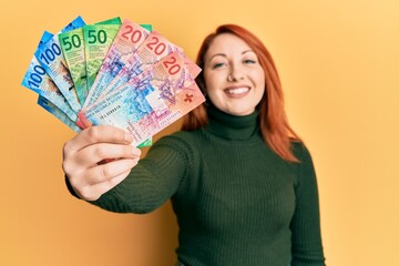 Canvas Print - Beautiful redhead woman holding swiss franc banknotes looking positive and happy standing and smiling with a confident smile showing teeth