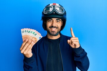 Young hispanic man wearing motorcycle helmet holding 10 euros smiling with an idea or question pointing finger with happy face, number one