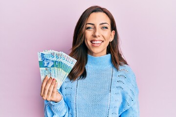 Poster - Young brunette woman holding hong kong 20 dollars banknotes looking positive and happy standing and smiling with a confident smile showing teeth