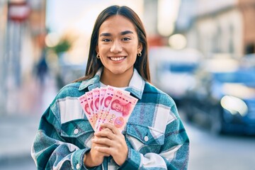 Sticker - Young latin girl smiling happy holding chinese yuan banknotes at the city.