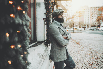 Portrait of a dapper bearded handsome African guy in a woolen cap, stylish eyeglasses, and elegant custom-made business costume, leaning against the wall decorated with Christmas trees and garlands