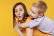 © ViDi Studio - Happy young woman have fun with cute child baby boy 5-6-7 years old in violet t-shirt stand behind hug kiss Mommy little kid son together isolated on yellow background studio Mother's Day love family