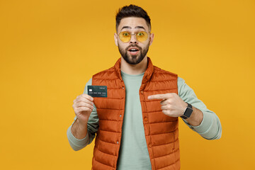 Wall Mural - Young surprised shocked amazed happy rich successful fun caucasian man 20s in orange vest mint sweatshirt glasses point index finger on credit bank card isolated on yellow background studio portrait