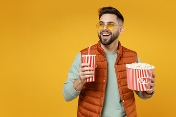 Poster - Young smiling happy cheerful fun caucasian man 20s in orange vest mint sweatshirt glasses holding popcorn takeaway bucket cup of soda cola watching film isolated on yellow background studio portrait