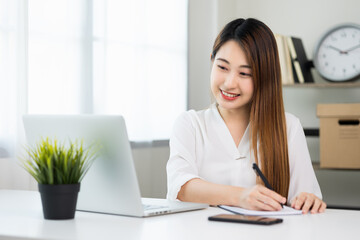 Young asian beautiful business woman working with laptop sitting at home and note on book. Smiling charming happy young female doing homework meeting conference with team at home.