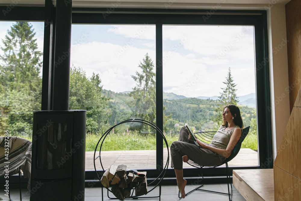 Beautiful stylish woman reading book, sitting on chair at fireplace on background of mountain hills - obrazy, fototapety, plakaty 