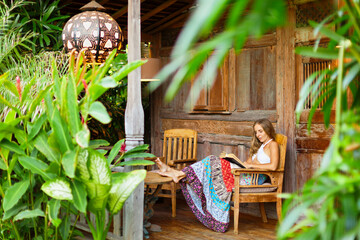 Attractive girl sit on open-air veranda of wood house with tropical garden view, read romance in paper book. Young woman relaxing in luxury villa on family summer vacation. Tropical island lifestyle.