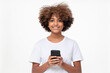 © Damir Khabirov - African american smiling teenage girl with afro hairstyle holding phone with both hands, chatting with friend, using social media app, isolated on gray background