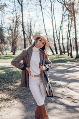 Wall Mural - Beautiful woman wearing stylish blazer, hat and boots holding purse in park. Spring female fashionable outfit.