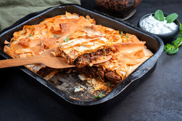 Wall Mural - Traditional spiced Moroccan lamb ragout with filo pie served as close-up in a rustic black backing dish on a board