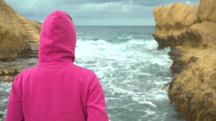 A young woman is standing in front of a cliff in the sea. The sea among the rocks. A woman stands with her back in a pink hoodie. Storm on the Mediterranean Sea.